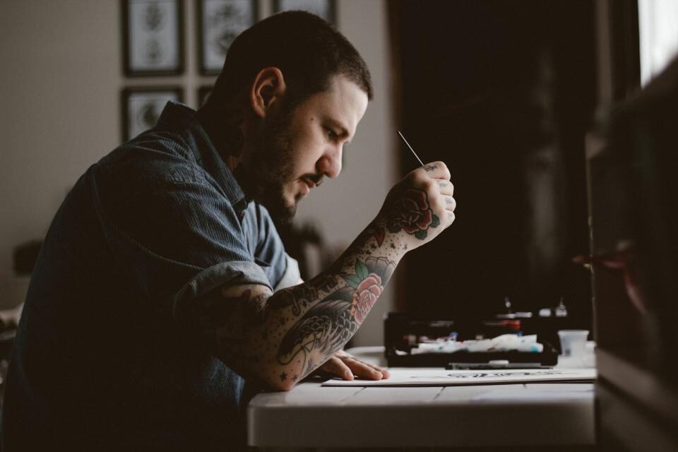 Tattooed artist at a desk drawing a design by window light